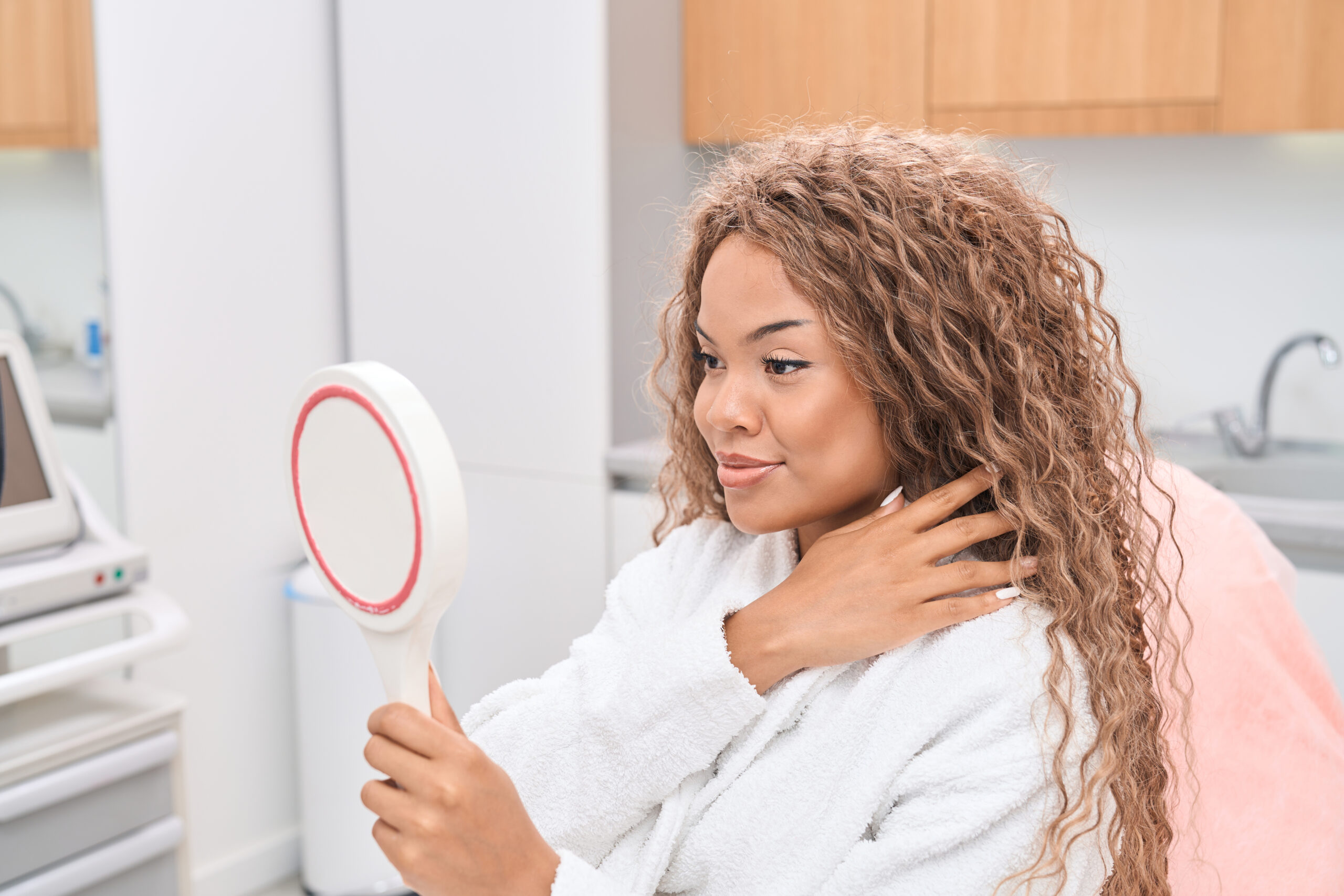 Smiling young woman in the office of a modern aesthetic medicine salon holding a round mirror in her hands